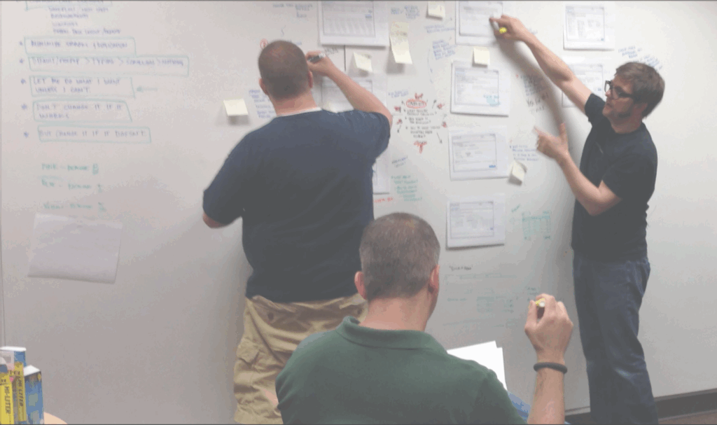 Derek Fricano leads a design review on a wall-sized whiteboard. He points at papers that are taped to the wall while another man writes on the whiteboard wall.